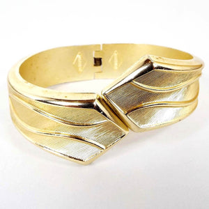 Front view of the Mid Century vintage Sarah Coventry hinged bangle. The bracelet is gold tone in color. It opens at the front and the ends are V shaped. There is a textured line design with shiny wavy lines over the top. It hinges in the back.