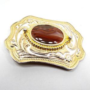 Top view of the retro vintage enameled belt buckle with agate gemstone. The metal is gold tone in color and has a curled leaf and floral design around the belt buckle. There is white enamel around the floral and leaf design. In the middle is a large oval agate gemstone cab that has has marbled clear, brown, and orange tones.