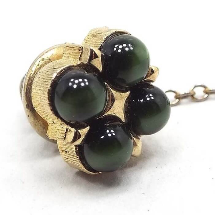 Angled enlarged front view of the Mid Century vintage lucite tie tack. the metal is gold tone in color. There are four domed round lucite cabs in olive green that have a glowy like appearance as you move around in the light. The clutch has a chain coming off of it.