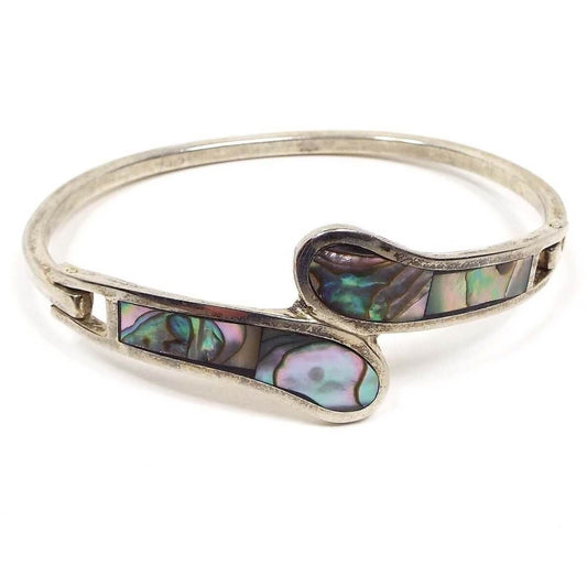 Front view of the retro vintage Taxco bypass hinged bangle bracelet. It is slightly darkened silver tone in color. The front has two rounded end areas that bypass one another with inlaid abalone shell.