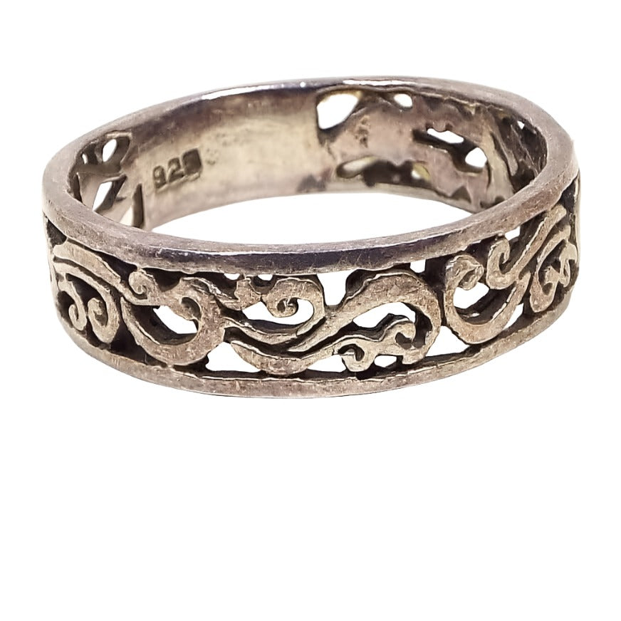 Angled front view of the sterling silver filigree band ring. It has an Art Nouveau style cut out design of curves and curls. The filigree goes almost all the way around the band except for a small closed smooth portion on the bottom of the ring. 925 is stamped on the inside of the band. The sterling is slightly darkened from age.
