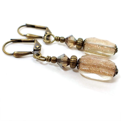 Angled side view of the handmade faceted glass drop earrings. The metal is antiqued brass in color. There are brown faceted glass crystals at the top. The bottom glass beads are faceted rectangle and are clear with fine copper color glitter inside them.