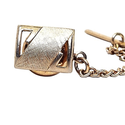 Enlarged front view of the Mid Century vintage Swank brushed matte gold tone tie tack. It is rectangle in shape and has a cut out corner area on each side diagonal from each other. There is a middle diagonal stripe area with textured brushed matte gold tone. There is a chain on the clutch back with a small bar on the end.