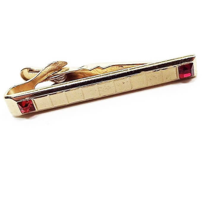Front view of the thin Mid Century vintage Swank rhinestone tie clip. It is gold tone in color and has a small square pattern across the front. There is a square deep red rhinestone on each end.
