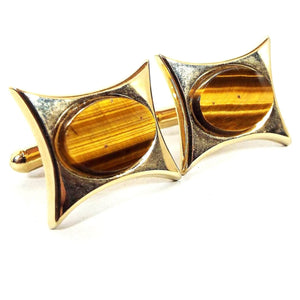 Angled front view of the Mid Century vintage gemstone cufflinks. They have a sort of rectangle shape with curved in sides leaving pinched angles at the corners. There are oval tiger's eye gemstone cabs in the middle with shades of yellow and brown.