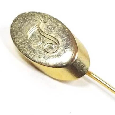 Enlarged view of the top of the Mid Century vintage initial stick pin. The metal is gold tone in color. There is an oval shape at the top with fancy script letter T engraved on it.
