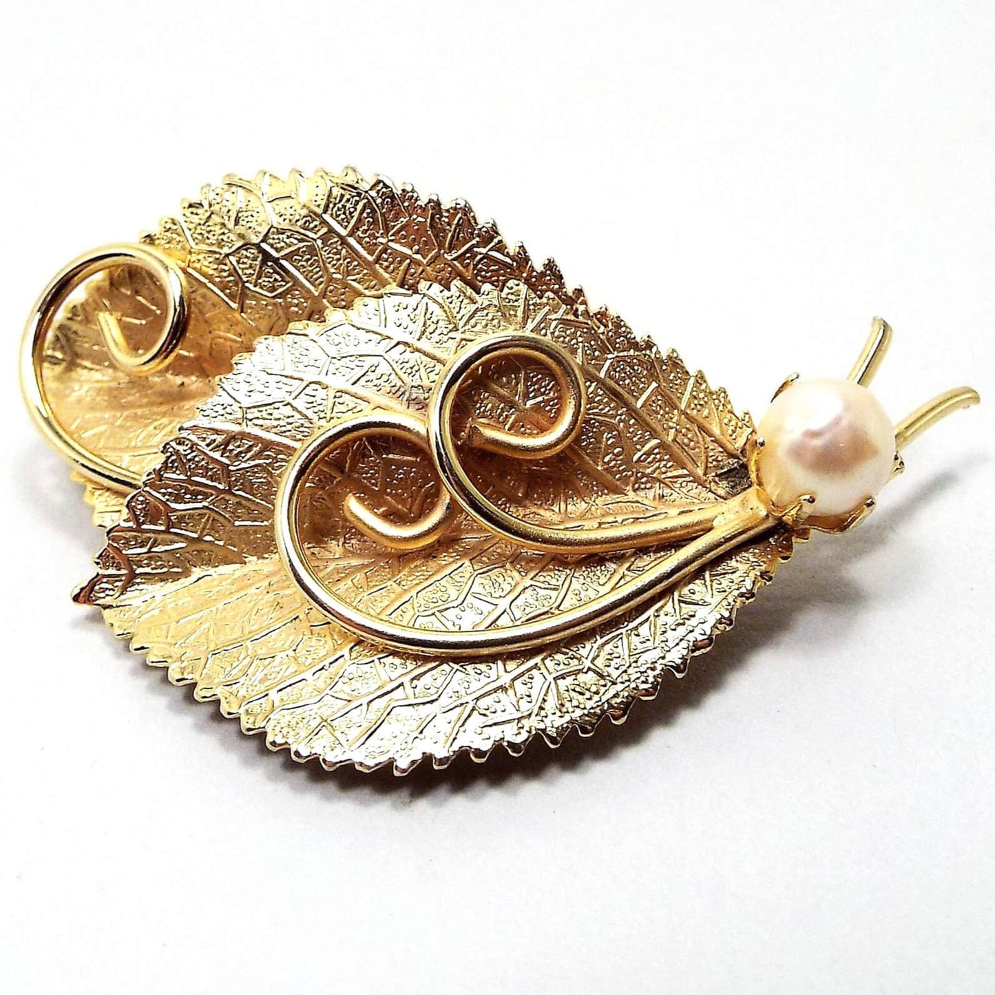 Front view of the retro vintage cultured pearl brooch. The metal is gold tone in color. It is designed like two textured leaves with stems and metal curls in the middle. At the bottom by the stems is a prong set round cultured pearl in a light off white color.