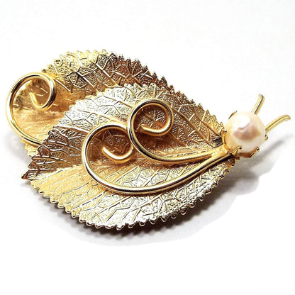 Front view of the retro vintage cultured pearl brooch. The metal is gold tone in color. It is designed like two textured leaves with stems and metal curls in the middle. At the bottom by the stems is a prong set round cultured pearl in a light off white color.