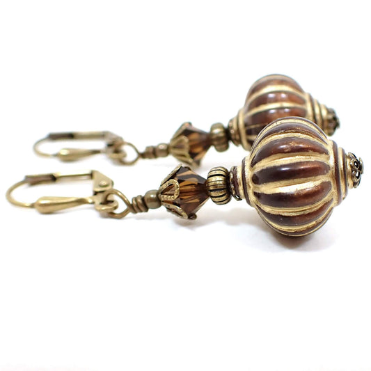 Side view of the handmade lantern drop earrings. The metal is antiqued brass in color. There are dark brown faceted glass crystal beads at the top and brown acrylic lantern shape beads at the bottom. The bottom beads have antiqued gold stripes painted on them.