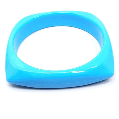 Angled view of the retro vintage plastic bangle bracelet. It is a bright blue in color and has a square shape with rounded corners. 