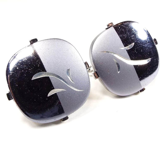 Front view of the Mid Century vintage Swank cufflinks. They are shaped like a square with rounded edges. The design is split in half with metallic gray on one side and a lighter matte gray on the other. There is an etched silver tone leaf design in the middle.