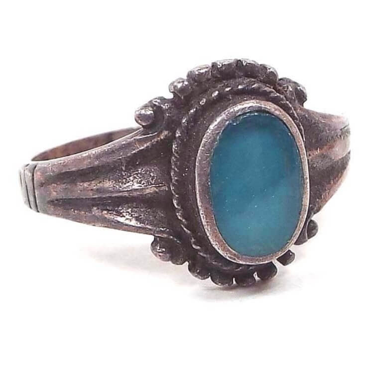 Angled front and side view of the retro vintage sterling silver chalcedony ring. The sterling is darkened in color from age to a gray color. The chalcedony gemstone cab on the top is oval and flush with the bezel. There is a braided rope design around the oval and curved sterling dot design on the top and bottom of the bezel area. The sides of the ring flares towards the bezel and tapers down to the band.