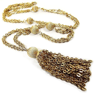 Angled view of the Mid Century vintage multi strand tassel necklace. The metal is gold tone in color. There are two strands of chain with a few larger round corrugated beads down the necklace. The bottom has a large tassel with numerous strands of chain. 
