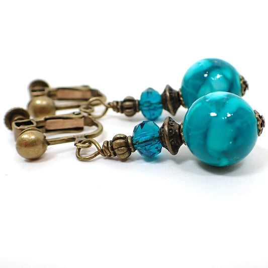 Side view of the handmade drop earrings. The metal is antiqued brass in color. There are teal blue faceted glass crystals at the top and marbled teal blue acrylic round beads at the bottom.