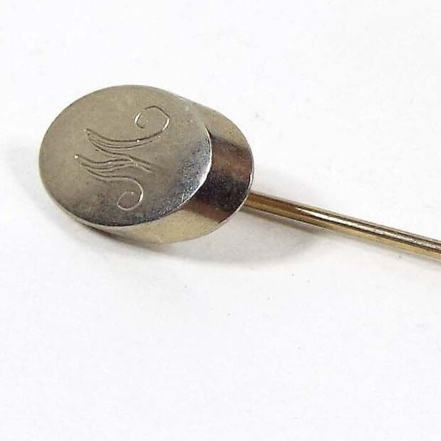 Enlarged view of the top of the Mid Century vintage initial stick pin. The metal is a light gold tone in color. The top has an oval part with the letter M engraved on it in fancy script. 
