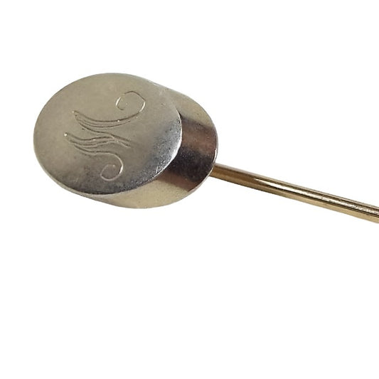 Enlarged view of the top of the Mid Century vintage initial stick pin. The metal is a light gold tone in color. The top has an oval part with the letter M engraved on it in fancy script. 