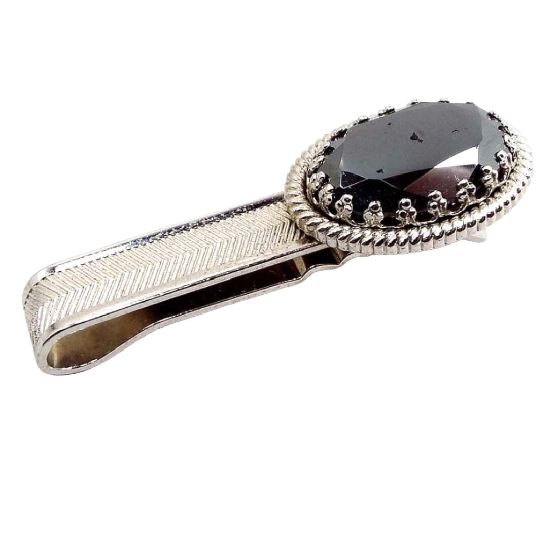 Angled front view of the Mid Century vintage faux hematite tie bar. The metal is silver tone in color. It is a slide on style tie bar with a large faceted oval glass imitation hematite cab at the end.