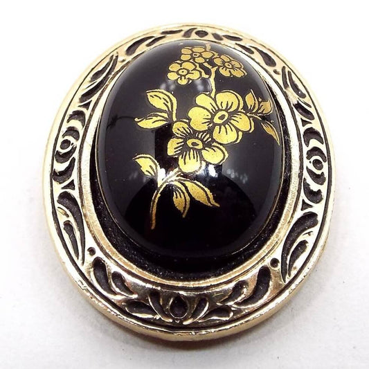 Front view of the West Germany Mid Century vintage floral scarf clip. It is oval in shape. There is a black glass cab on the front with a metallic gold painted flower design. The outer edge is plastic with an indented black design and metallic gold color.
