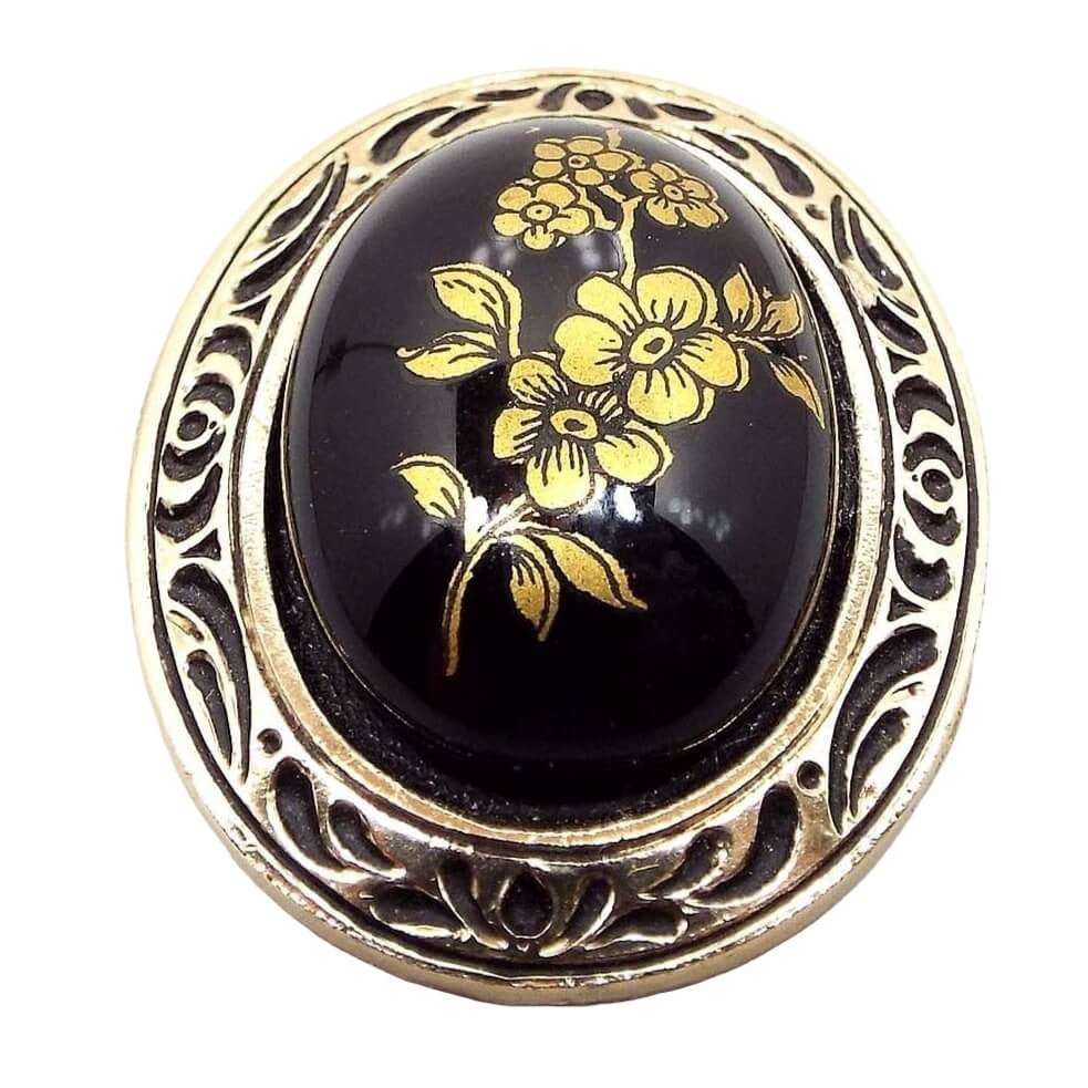 Front view of the West Germany Mid Century vintage floral scarf clip. It is oval in shape. There is a black glass cab on the front with a metallic gold painted flower design. The outer edge is plastic with an indented black design and metallic gold color.
