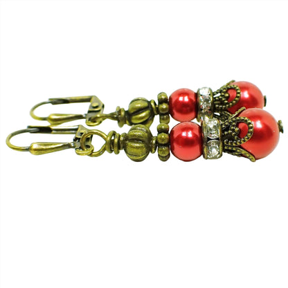 Side view of the handmade glass beaded earrings with rhinestones. The metal is antiqued brass in color. The earrings are beaded with a round metallic red coated glass bead, a rhinestone spacer bead, and then another larger round metallic red glass bead. 