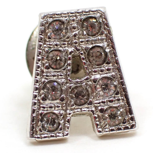 Enlarged front view of the retro vintage rhinestone initial tie tack. It is silver tone in color and shaped like a letter A.  There are round clear glass crystal rhinestones along the entire front of the tie tack. There is a very tiny green spot of verdigris next to one of the rhinestones that can be seen under magnification.