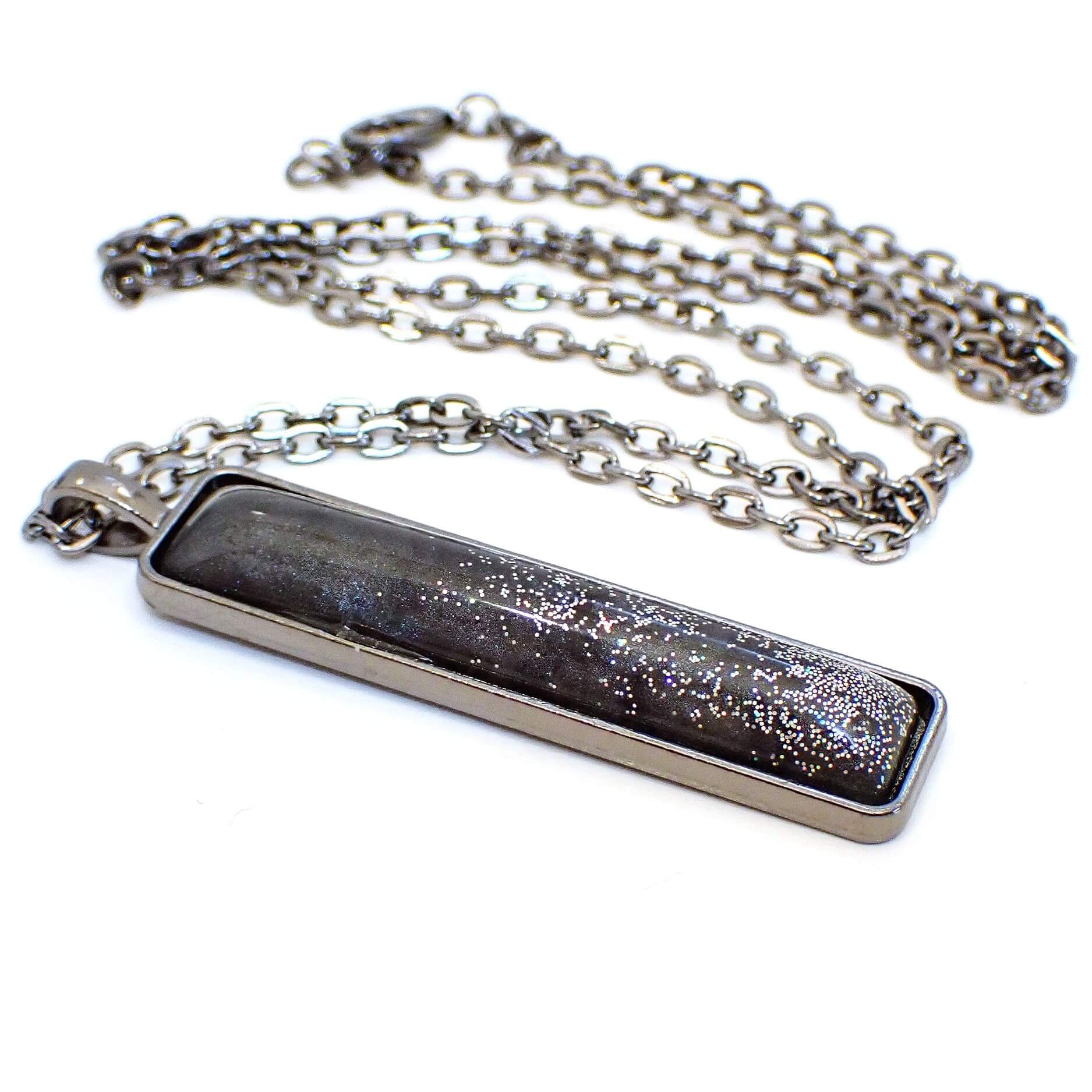 Angled view of the handmade resin bar pendant necklace. The metal is gunmetal gray in color. The long rectangle pendant has rounded edges and a handmade resin cab on the front. The resin cab has pearly dark gray resin with tiny flecks of holographic glitter at the bottom half of the pendant.