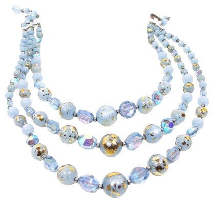 Front view of the Mid Century vintage multi strand necklace. There are three strands of light blue beads. There are round moonglow lucite and round lucite beads with metallic gold color splatter paint on them. In between here and there are AB blue crystal beads in a faceted sauce shape. The end clasp and other metal parts are silver tone in color.