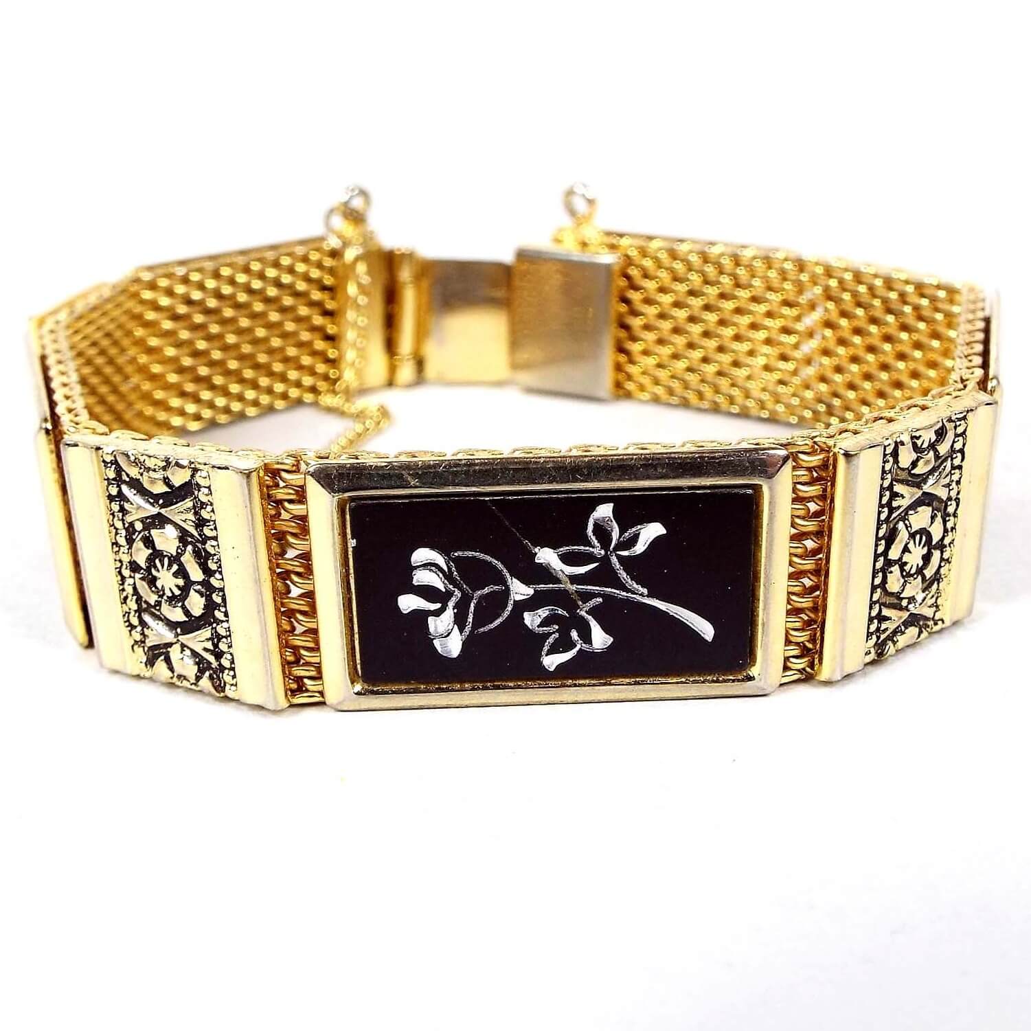 Front view of the Mid Century vintage Damascene style floral bracelet. It has a wide gold tone mesh setting with rectangle links. The smaller links have a flower design with a black background. The larger links are black with a silver tone color etched flower on them. there is a box clasp and a safety chain at the end.