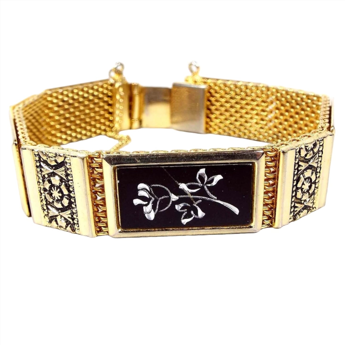 Front view of the Mid Century vintage Damascene style floral bracelet. It has a wide gold tone mesh setting with rectangle links. The smaller links have a flower design with a black background. The larger links are black with a silver tone color etched flower on them. there is a box clasp and a safety chain at the end.