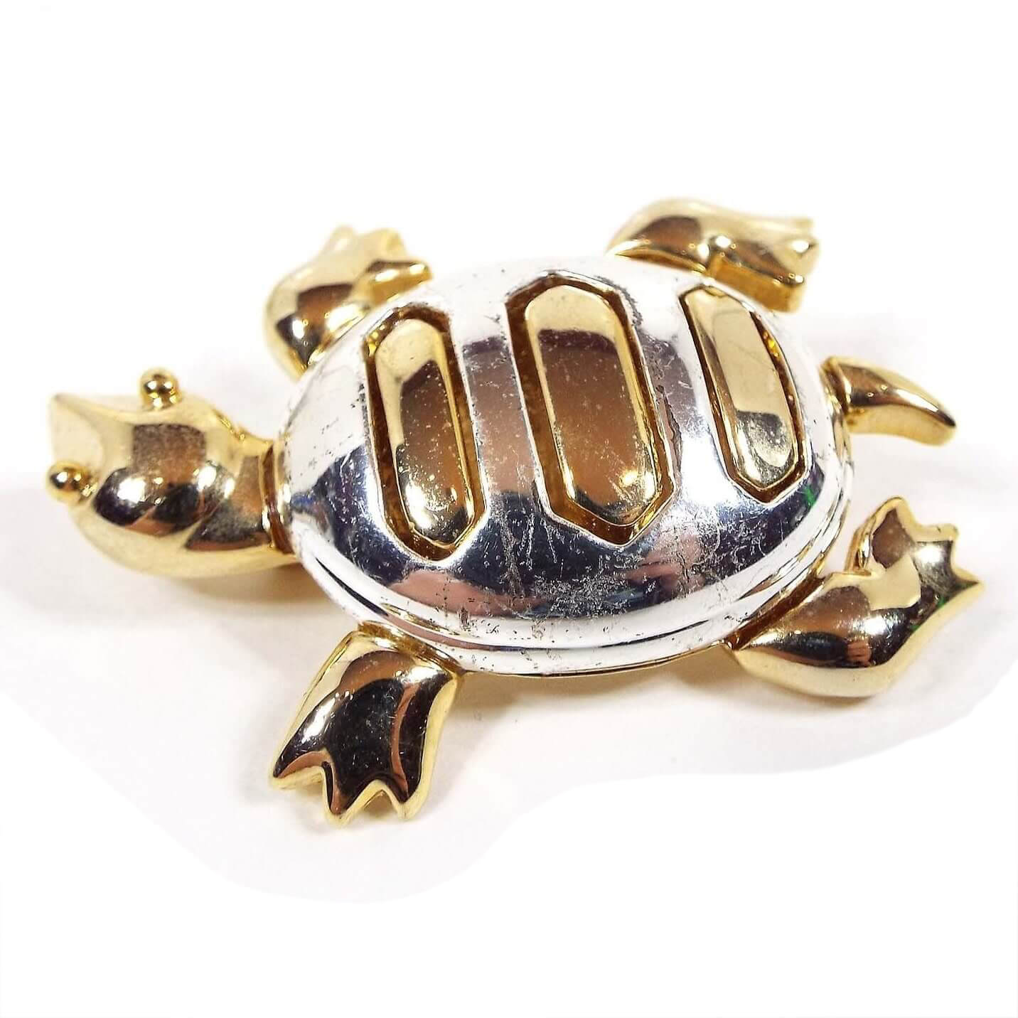 Top view of the Allison Reed retro vintage turtle brooch pin. The turtle appears to be on the move with his legs bent and his head turned to the right. His head, legs, tail, and stripes on top are gold tone in color and his shell is silver tone in color.