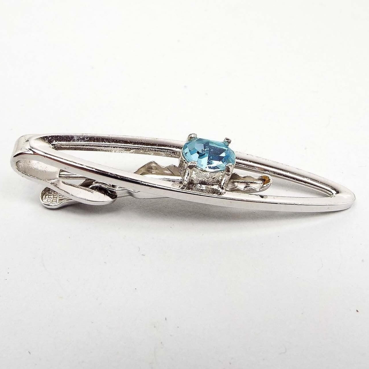 Angled front view of the Mid Century vintage Dante rhinestone tie clip. The metal is silver tone in color. It has an open long oval design with a light blue oval rhinestone in the middle. The rhinestone is prong set. 