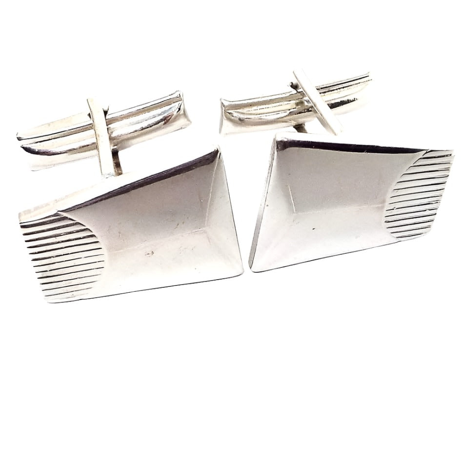 Angled front view of the Sarah Coventry Mid Century vintage cufflinks. They are silver tone in color. They have a trapezoid shape with lines engraved on one side.