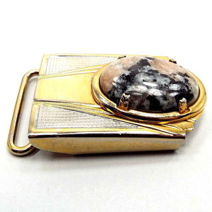 Angled front and side view of the retro vintage gemstone belt buckle. It is mainly gold tone in color with some silver tone areas on the top edge. There is a loop on the back and an oval rhodonite gemstone cab on the front in shades of gray and pink.