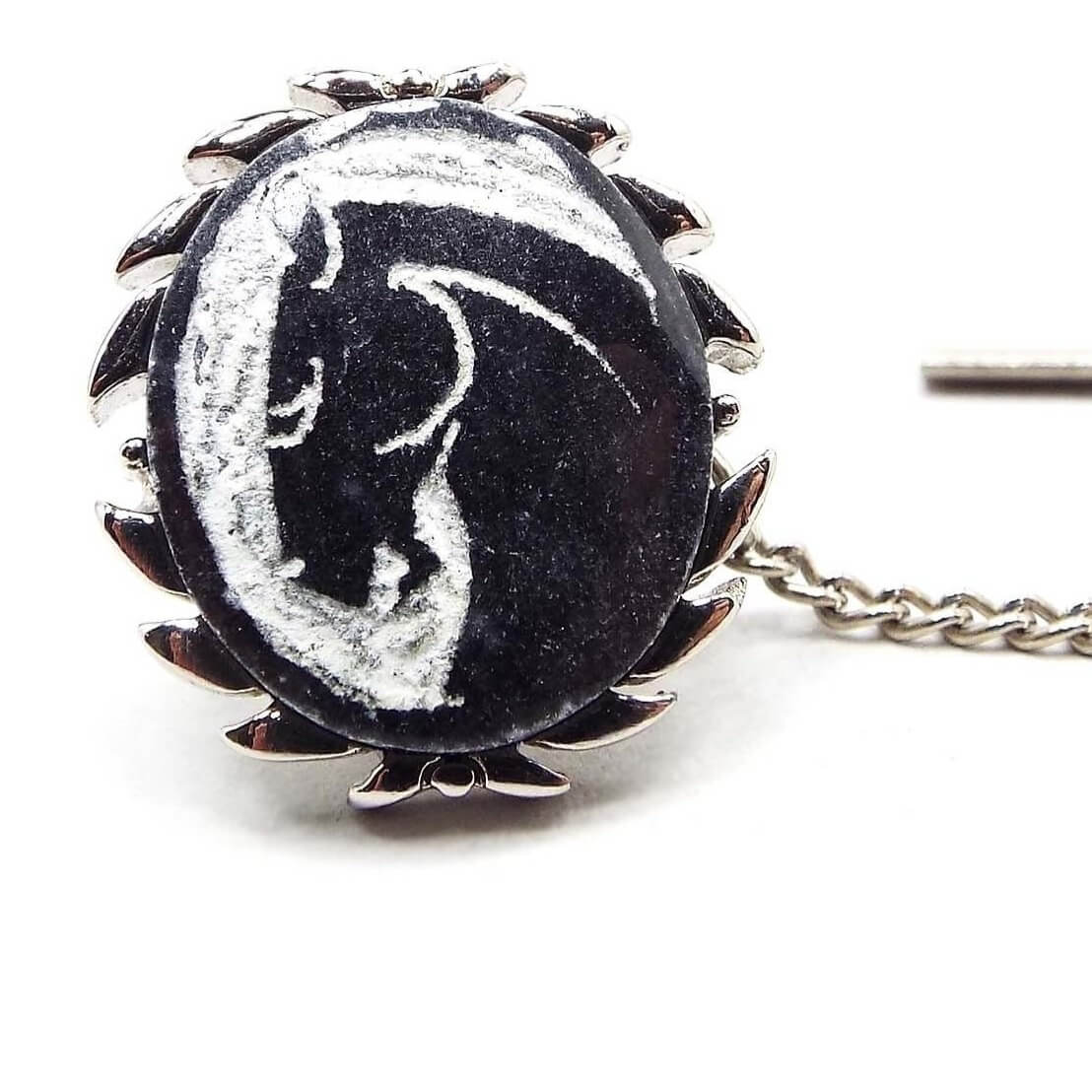 Enlarged front view of the retro vintage Swank carved stone tie tack. The metal is silver tone in color and has a leaf like curved design around the edge. The stone cab is black with white showing what's been carved away and has a horse head cameo.