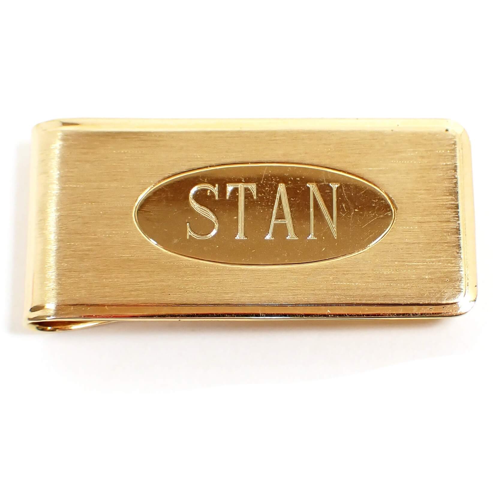 Front view of the retro vintage name money clip. The metal is gold tone in color. The front has a matte brushed finish with a shiny oval in the middle. The name Stan is engraved inside the oval.