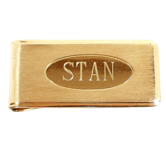 Front view of the retro vintage name money clip. The metal is gold tone in color. The front has a matte brushed finish with a shiny oval in the middle. The name Stan is engraved inside the oval.