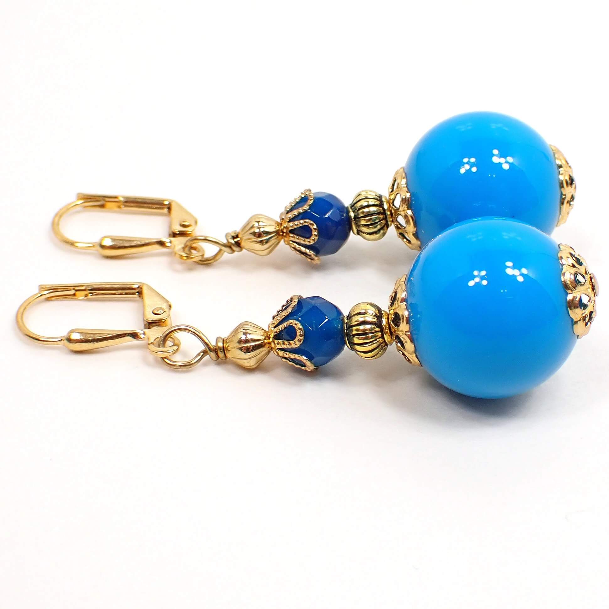 Side view of the handmade gumball drop earrings. The metal is gold plated in color. There is a new faceted glass bead in dark teal blue at the top and a bright blue vintage lucite round bead at the bottom.