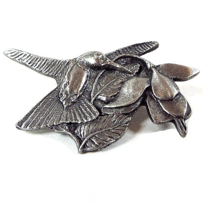 Front view of the retro vintage Birds and Blooms Premiere Edition brooch pin. It is made of pewter and gray in color. There is a flying hummingbird flying next to a Fuchsia bloom that hangs down on the right side of the brooch.