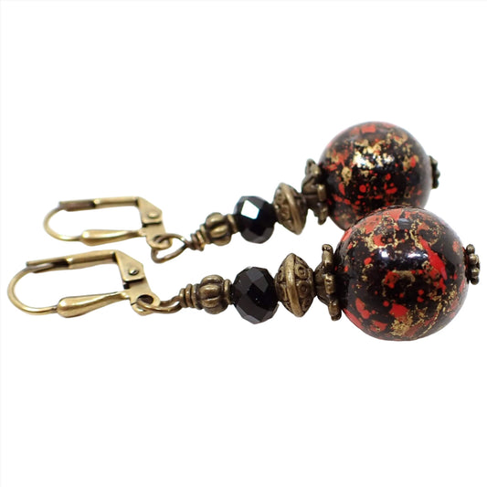 Side view of the handmade round drop earrings. The metal is antiqued brass in color. There are faceted glass crystal black beads on the top. the bottom beads are round lucite beads with marbled black, red, and metallic antiqued gold color. 