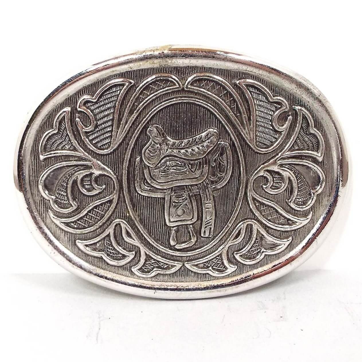 Front view of the retro vintage Avon belt buckle. It is silver tone in color with a light gray background. There is a horse saddle in the middle and a floral like pattern around the edge.