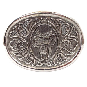 Front view of the retro vintage Avon belt buckle. It is silver tone in color with a light gray background. There is a horse saddle in the middle and a floral like pattern around the edge.