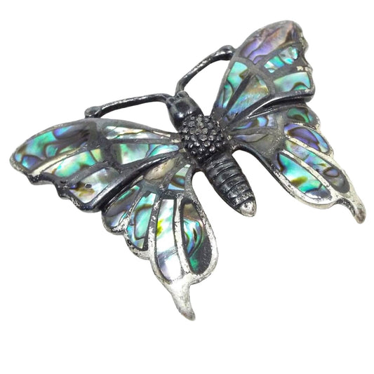 Front view of the retro vintage Southwestern Boho butterfly brooch pin. The sterling silver is very darkened in color to a really dark gray and black in some areas. The brooch is shaped like a butterfly with a textured body. The wings have areas on inlaid abalone shell that have various flashes of color as you move around in the light.