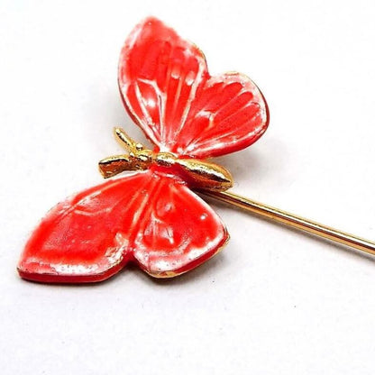Magnified view of the top part of the retro vintage enameled butterfly stick pin. The metal is gold tone in color. The butterfly is a bright coral pink in color with a gold tone body. 
