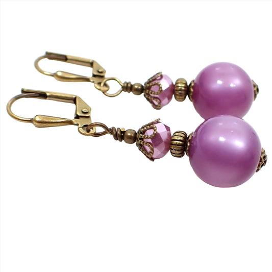 Side view of the handmade lucite drop earrings. The metal is antiqued brass in color. There are faceted light purple glass beads at the top. The bottom beads are round moonglow lucite beads in a lilac purple color. The lucite beads have a glowy effect as you move around in the light.