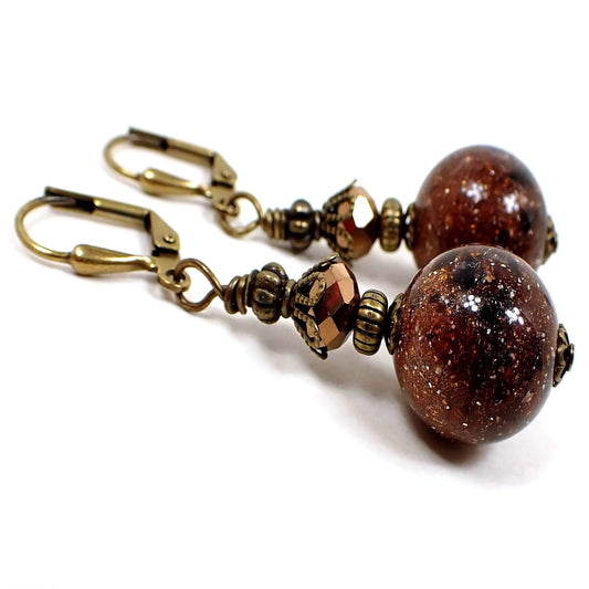 Angled side view of the handmade sparkly brown lucite galaxy earrings. The metal is antiqued brass in color. It has a faceted glass metallic brown bead at the top and a round lucite bead at the bottom that has tiny sparkles and swirls of brown and black that resembles a galaxy like appearance.