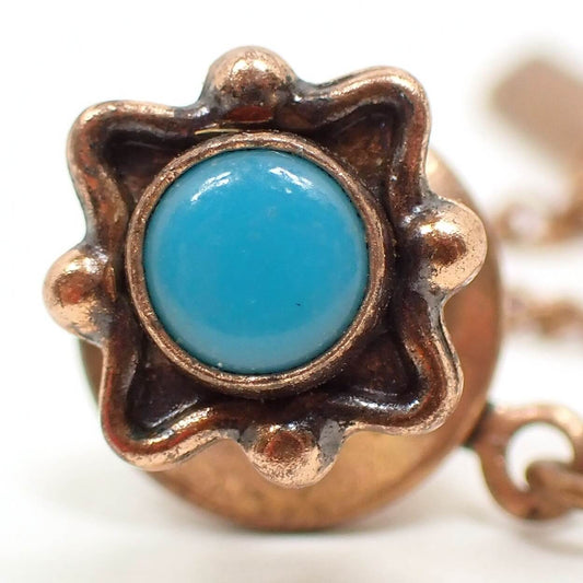 Enlarged front view of the Southwestern style retro vintage tie tack. The metal is antiqued copper tone plated in color. It is shaped like a square with pinched in sides that have round spheres in the middle of each side. There is a flat round plastic cab on the front that is turquoise blue in color.