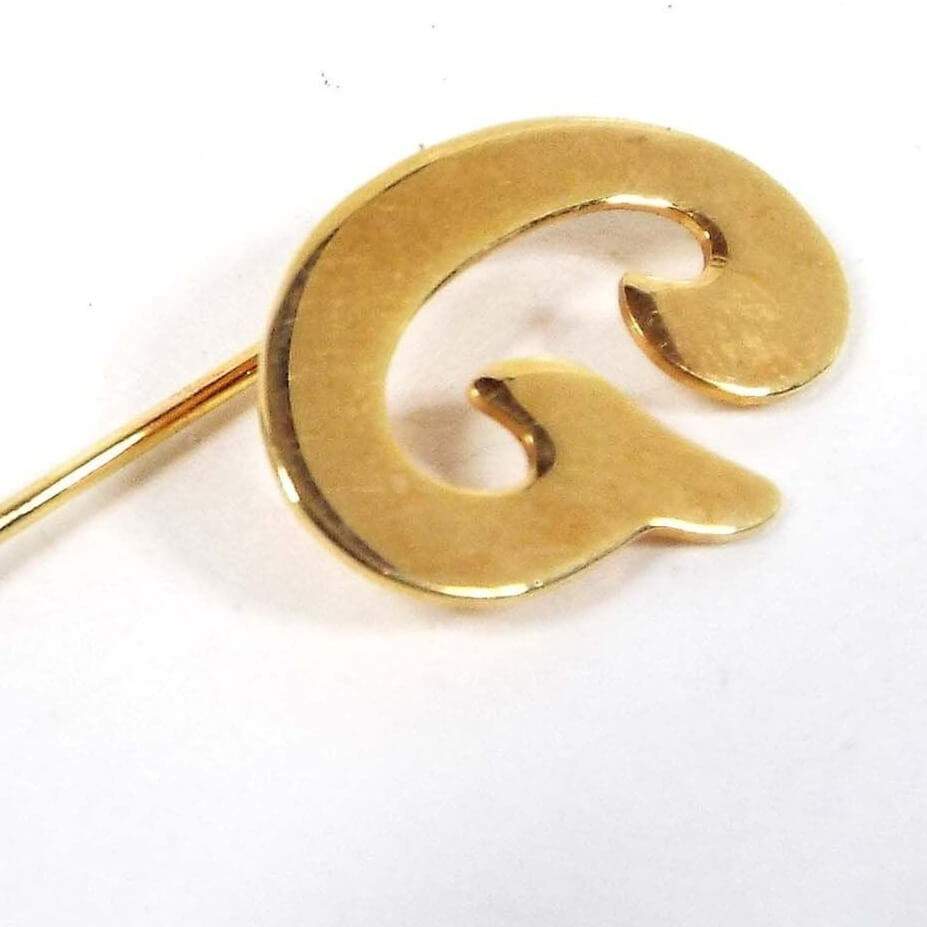 Enlarged top view of the retro vintage initial stick pin. The metal is gold tone in color. There is a curvy letter G at the top.