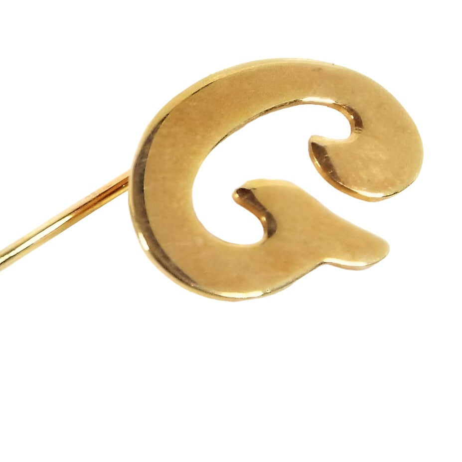 Enlarged top view of the retro vintage initial stick pin. The metal is gold tone in color. There is a curvy letter G at the top.