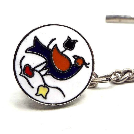 Angled front view of the retro vintage enameled tie tack. It is round in shape with silver tone plated color metal. The front is enameled white with a blue and red pheasant bird design with blue and yellow flowers and a red heart on the bottom.