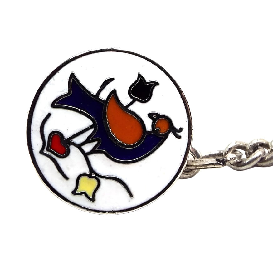 Angled front view of the retro vintage enameled tie tack. It is round in shape with silver tone plated color metal. The front is enameled white with a blue and red pheasant bird design with blue and yellow flowers and a red heart on the bottom.